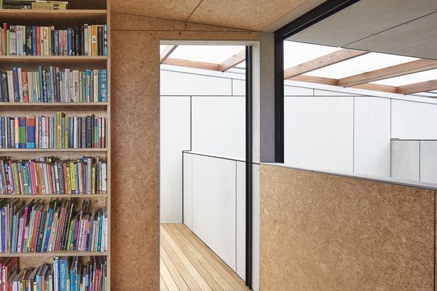 Compact contemporary interior with a full-height built-in bookshelf filled with books on the left, opening toward a bright minimalist corridor or workspace. The space features exposed cork or wood-fibre board wall and ceiling panels, pale timber floorboards, white built-in wall panels, and black-framed internal glazing or partitioning. Exposed timber roof beams are visible through the opening, creating a warm yet highly functional architectural interior.