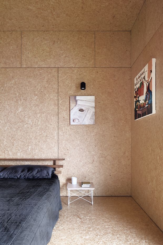 A compact bedroom with warm, textured natural-fibre wall and ceiling panels, matching flooring, a low timber bed with dark charcoal bedding, and a small white metal bedside table. Minimal artwork and a black wall-mounted reading light add restrained visual accents. The room has a simple, calm, minimalist composition with full-height wall panelling and a standard ceiling.