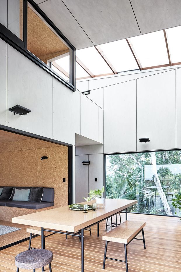 A bright, double-height open-plan dining and living area with a long light-toned dining table, benches, stools, and a built-in sofa. The room features exposed structural timber, pale white wall and ceiling panels, dark metal window and lighting frames, a large glazed opening to lush vegetation and water, and warm timber flooring. A high-level opening and translucent roof glazing bring in abundant natural light, creating a modern, airy interior.