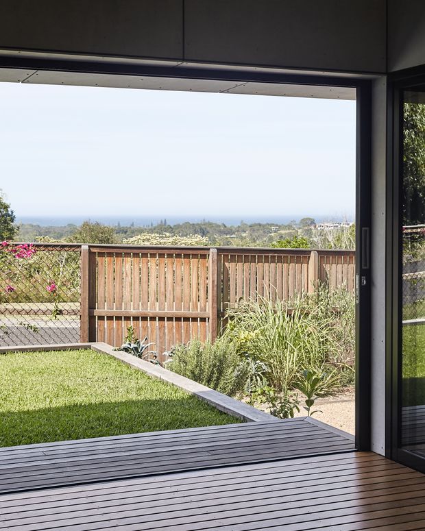 View from inside a modern home through a wide open glazed opening onto a timber deck and landscaped backyard. The garden includes lawn, planted garden beds and a timber privacy fence, with a distant coastal or ocean horizon visible beyond the suburban landscape. Dark-framed glazing and a dark timber-lined ceiling create a strong contemporary indoor–outdoor connection.