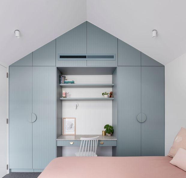 A compact bedroom with a built-in muted blue-grey storage and study wall designed around a pitched-roof profile. The cabinetry includes tall doors with circular handles, open display shelving, a desk with drawers, and an integrated upper air-conditioning vent. A pale pink bed occupies the foreground, with a white spindle-back chair at the desk and sparse decorative accessories. The room has white walls and ceiling, soft natural light, and a calm, contemporary built-in aesthetic.