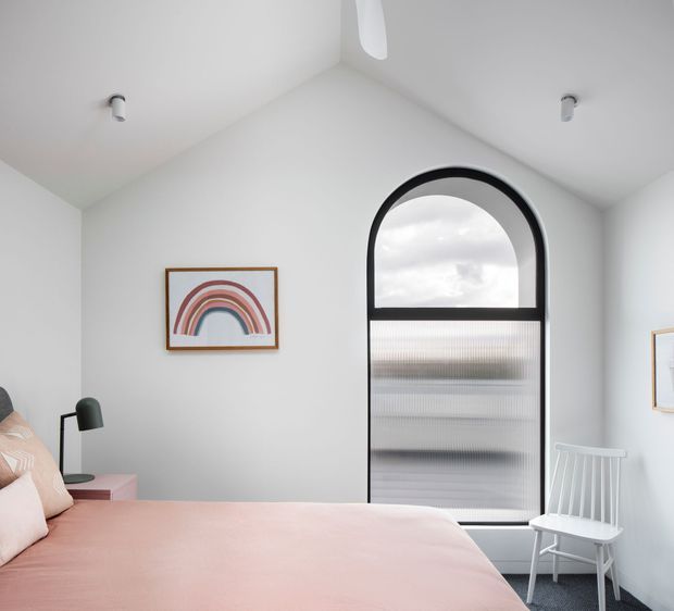 Bright, minimalist bedroom with a soft pink bedspread, white walls, a black bedside lamp, simple rainbow artwork, and a distinctive black-framed arched window or door with ribbed translucent glazing. A white rocking chair sits near the opening, and the ceiling has a simple pitched form with recessed spotlights.