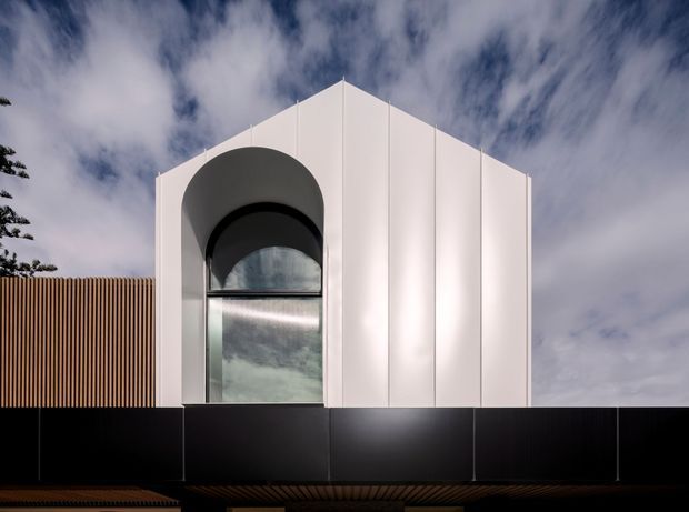 Front exterior view of a sculptural modern building facade with a crisp white gabled form, tall recessed arched window, and dark horizontal lower volume. Vertical timber slat screening is visible to the left, set against a dramatic cloudy sky.