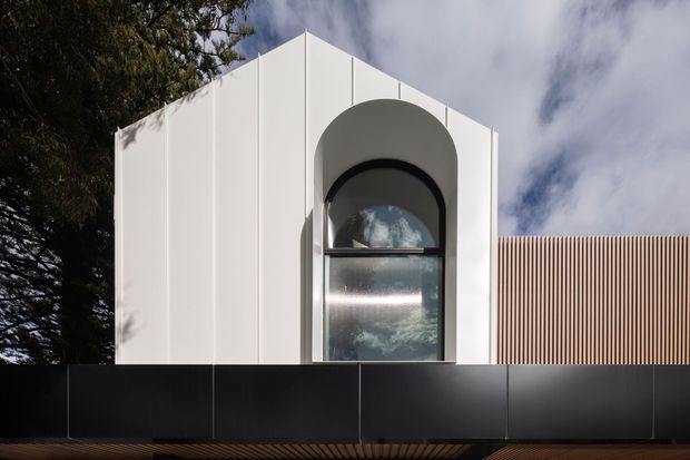 Contemporary white geometric building facade with a prominent tall arched window, black lower wall panels, and a vertical timber-toned slatted screen extending to the right. The image is an exterior architectural view framed by trees and sky.
