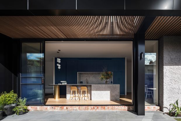 A modern indoor-outdoor kitchen viewed from a paved exterior terrace. The kitchen has a dark blue feature wall, large stone island with timber stools, timber flooring, expansive black-framed glazing, and a slatted timber ceiling creating a sheltered transition between exterior and interior. Potted plants and neighbouring structures are visible at the edges.