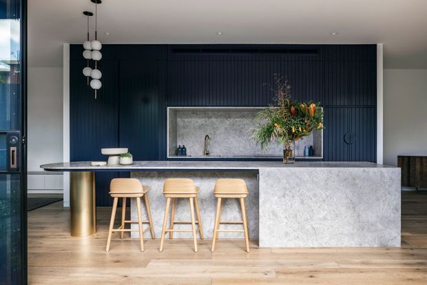 Contemporary kitchen with a large stone island and integrated breakfast bar, three timber bar stools, dark vertically ribbed cabinetry, a stone backsplash niche, pendant lighting, and timber flooring. Full-height glazing is visible at the left, with a dark timber and stone material palette.