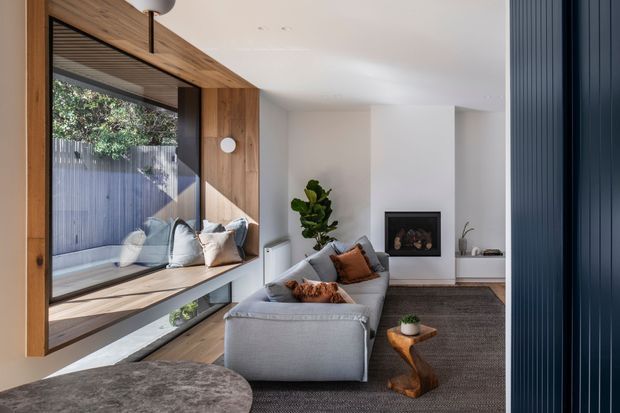 A contemporary living room with a large built-in timber-framed window seat and expansive glazing on the left, a grey sectional sofa, dark textured rug, white walls, and a recessed fireplace. A dark blue vertical-panel feature wall frames the right side, while a small sculptural timber side table and indoor plant add warmth.