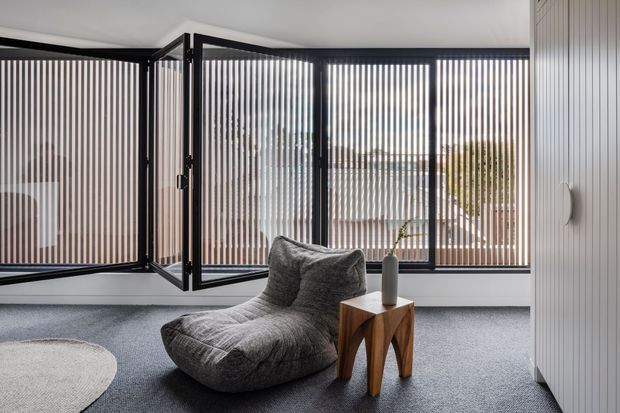 A minimalist sitting area with a sculptural grey lounge chair and small timber side table, set against full-height black-framed glazing with vertical privacy screening. White vertically panelled walls, grey carpet, and muted natural tones create a calm, contemporary interior.