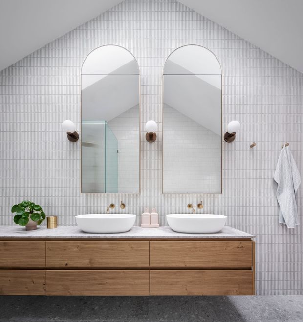 A refined double-vanity bathroom with a long timber floating vanity, two white vessel basins, brushed brass tapware and sconces, and matching arched mirrors. The walls are covered in fine vertical white tiles, with a glass shower enclosure reflected in the left mirror. Natural timber cabinetry and subtle greenery add warmth to the otherwise bright, minimalist interior.