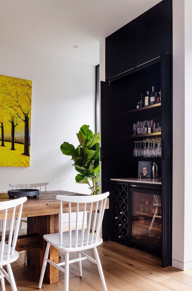 Bright dining area featuring a rustic timber dining table and white spindle-back chairs, beside a tall dark built-in drinks cabinet with open shelving, wine-glass storage, bottle display, wine racks and a glass-front beverage cooler. White walls, timber flooring, a leafy indoor plant and a vivid yellow tree artwork add warmth and contrast.