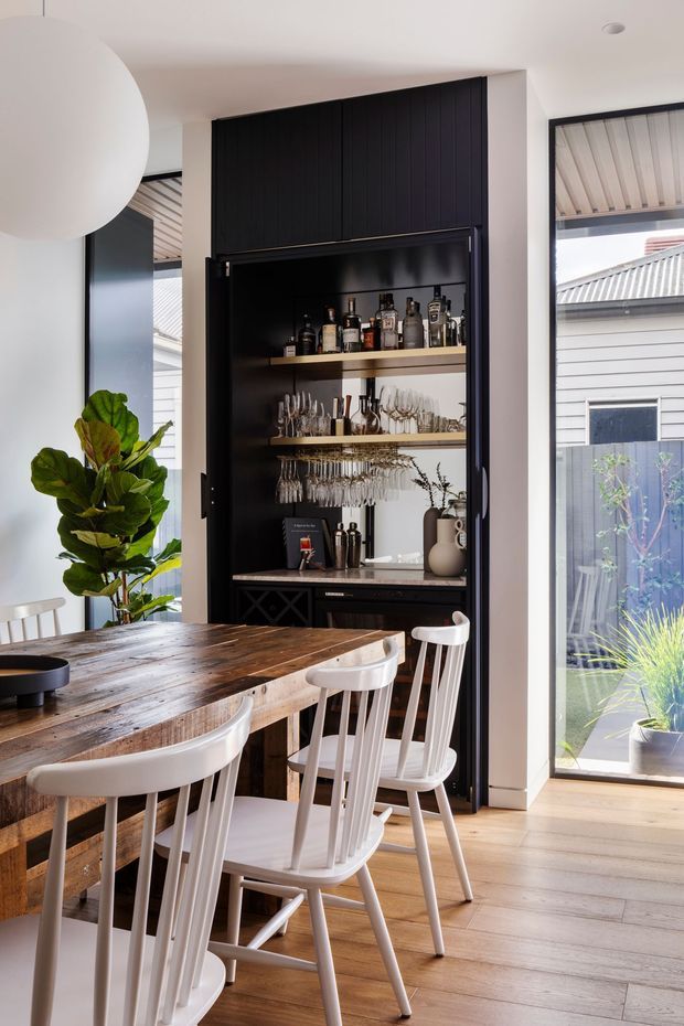 A bright dining area with a large rustic timber dining table and white spindle-back chairs. A dramatic black built-in home bar with open shelving, bottles, glassware, and a concealed lower cabinet forms the feature wall. Full-height windows provide abundant natural light and views to an adjacent exterior, while a leafy indoor plant and oversized spherical pendant add softness.