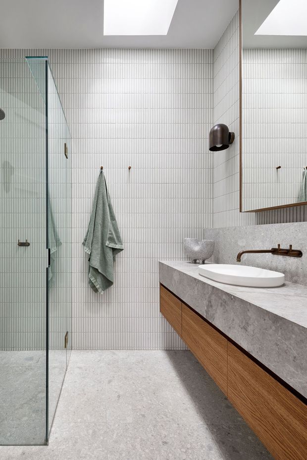Contemporary bathroom with a walk-in glass shower, full-height fine-format white wall tiles, pale stone-look floor and benchtop, a timber floating vanity, vessel basin, aged bronze wall-mounted tapware, and a large mirrored cabinet. A skylight provides bright natural illumination.