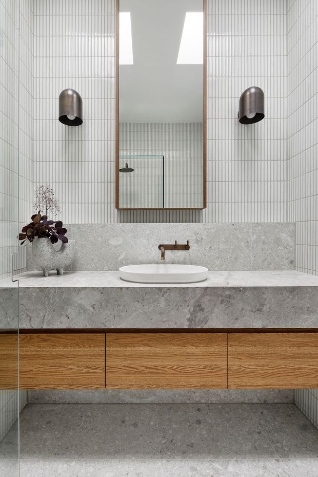 A refined contemporary bathroom vanity featuring a broad grey natural-stone benchtop and integrated backsplash, a white vessel basin, warm timber floating cabinetry, and a brushed bronze wall-mounted tap. The walls are clad in narrow vertically ribbed white tiles, complemented by a large timber-framed mirror, cylindrical metal wall sconces, and grey stone flooring.