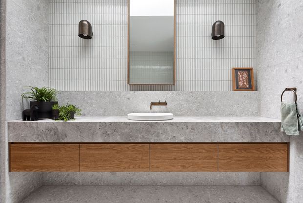 A refined contemporary bathroom vanity with a long floating timber drawer unit, pale grey stone benchtop and matching wall finish, a white vessel basin, brushed metal wall-mounted tap, large central mirror, vertically ribbed light tile backsplash, and dark wall sconces. Small plants, framed artwork, and a towel add restrained decorative accents.