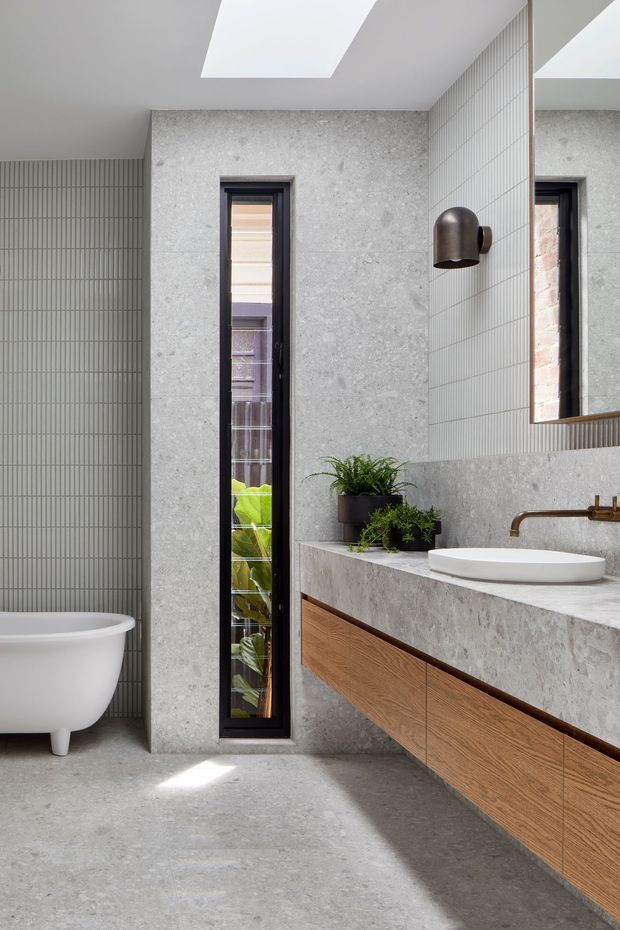 Contemporary bathroom with a freestanding white bathtub, vertically tiled walls, grey stone-look surfaces, a floating timber vanity with integrated stone benchtop, vessel basin and bronze tapware. A narrow window, skylight and large mirror bring in natural light, while potted greenery adds a natural accent.