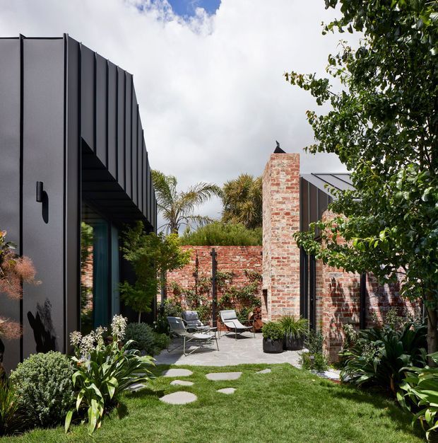 A landscaped courtyard between contemporary dark-clad and exposed-brick building volumes. The space includes a paved patio with outdoor lounge chairs, a brick chimney or feature wall, lush planting, lawn, and stepping stones. Vertical dark metal cladding and large glazing create a strong modern architectural contrast with the textured brickwork.