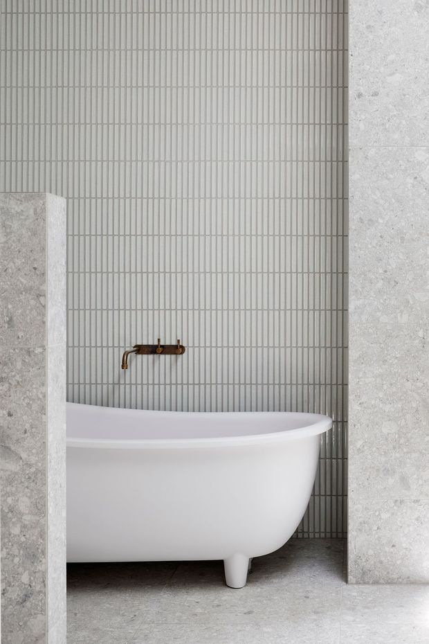 A refined bathroom detail featuring a white freestanding bathtub with a compact bronze wall-mounted tap, set against vertically ribbed light-grey tiles. Large pale stone-look wall slabs frame the bath, while matching grey stone flooring creates a calm, cohesive palette.