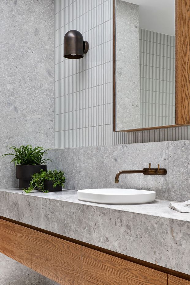 A refined contemporary bathroom vanity with a floating warm timber cabinet, pale grey stone wall and benchtop surfaces, a white vessel basin, and a wall-mounted aged bronze tap. Vertical ribbed wall tiles, a bronze wall sconce, and a large timber-framed mirror add texture and contrast. Small potted plants provide greenery.