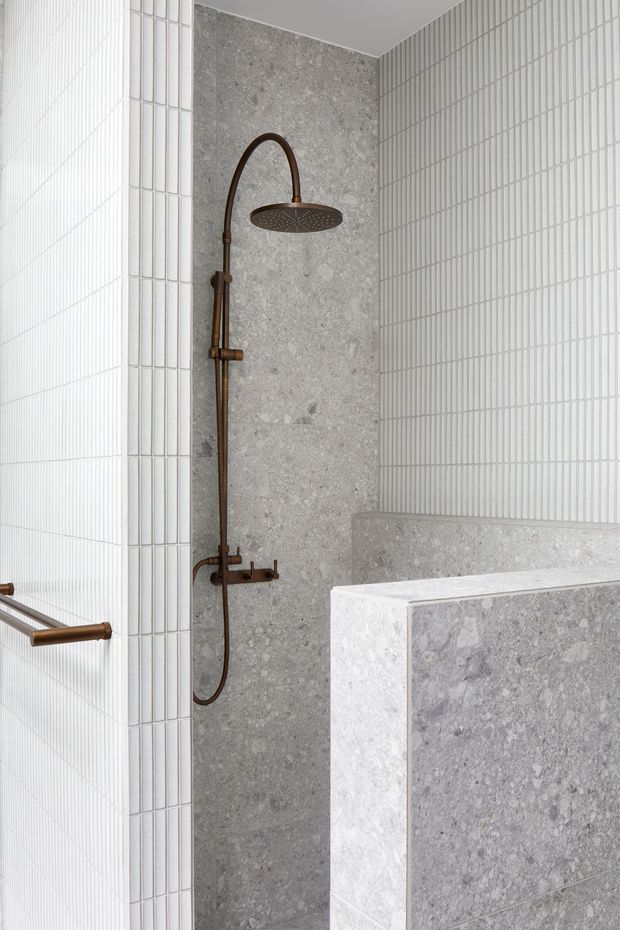 A refined walk-in shower featuring a curved bronze shower set with rainfall head, grey natural-stone wall and bench surfaces, and white vertically ribbed ceramic wall tiles. The partially enclosed shower has clean contemporary detailing and a light, neutral palette.