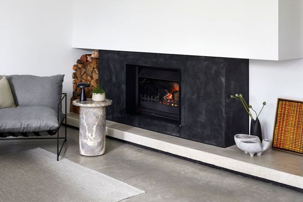 Contemporary living room centered on a built-in fireplace with a dark textured surround and broad white mantel. A grey lounge chair, sculptural stone side table, stacked firewood, ceramic vessels, artwork, and a neutral rug create a refined, monochromatic setting.