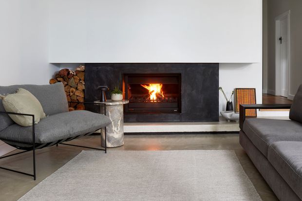 A refined living room centered on a built-in wood-burning fireplace with a dark stone or concrete surround and white overmantel. The room features a grey upholstered lounge chair and sofa, a large neutral rug, a slim black side table, firewood storage, and restrained contemporary furnishings.