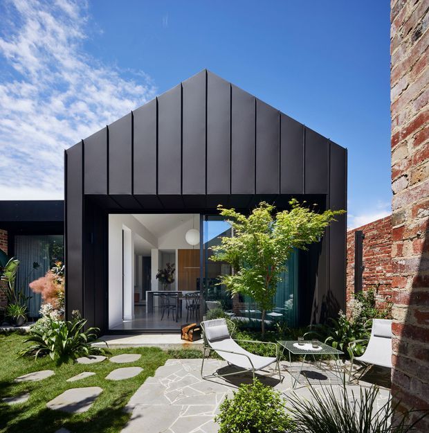 A contemporary black-clad gable-front extension opens onto a landscaped courtyard with large glazed doors, outdoor lounge furniture, stepping stones, planting, and a contrasting exposed brick wall at the right edge. The facade has vertically articulated dark metal panels and a crisp geometric form.