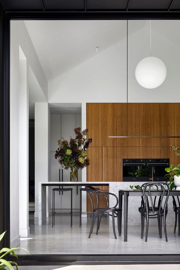 A refined open-plan kitchen and dining area viewed through large black-framed glazing. The space features full-height warm timber cabinetry with integrated black appliances, a pale stone island and dining table, black bentwood-style chairs and stools, polished light flooring, white sculptural walls, and a large spherical pendant light. Indoor plants and floral arrangements add natural accents.