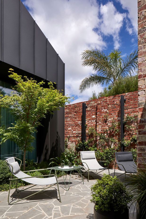 A landscaped urban courtyard terrace with a contemporary dark-clad building, exposed red-brick boundary walls, and pale irregular stone paving. Outdoor lounge chairs surround a small round table, with palms, shrubs, and climbing greenery adding a lush garden atmosphere. A tall brick wall and contrasting black vertical supports frame the space beneath a bright, partly cloudy sky.