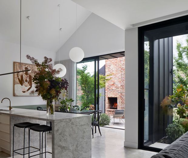 A bright, contemporary open-plan kitchen and dining area with a large stone island, black bar stools, sculptural pendant lights, and black-framed glazing. The space opens to a brick outdoor courtyard with a fireplace, while a black-framed glazed door and indoor greenery add strong indoor-outdoor connection.