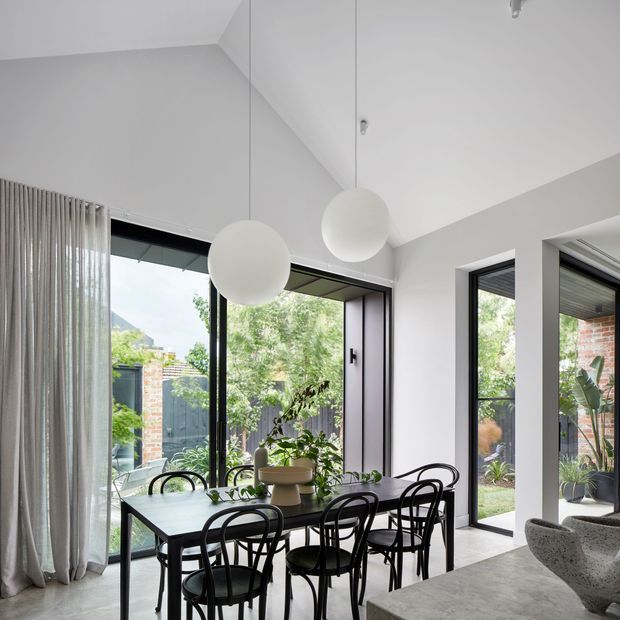 Bright contemporary dining area with a black dining table and curved black chairs beneath two large spherical pendant lights. Full-height black-framed glazing opens the room visually to a landscaped courtyard, while white walls, a high angled ceiling and sheer floor-to-ceiling curtains create an airy feel. A partial living area is visible at right.