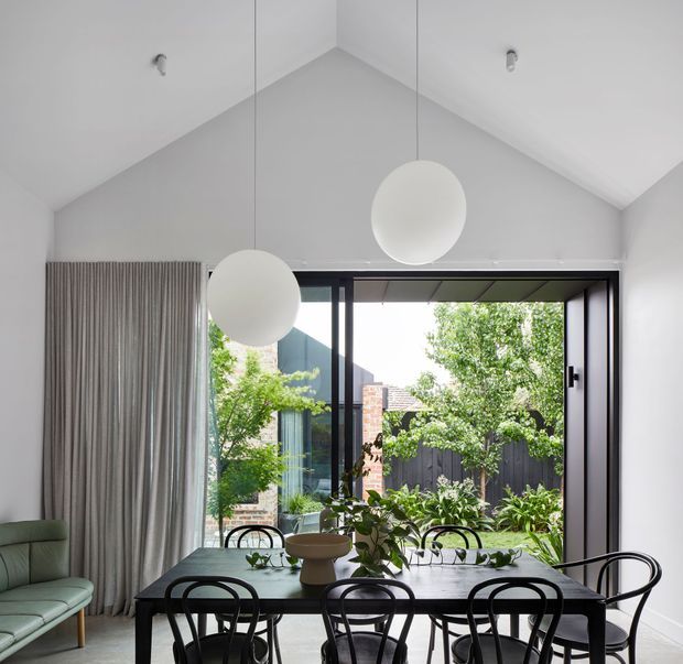 A bright, contemporary dining area with a long dark dining table and black bentwood chairs beneath two oversized spherical pendant lights. Full-height black-framed glazing opens to a leafy courtyard, while tall grey curtains and a steeply pitched white ceiling create an airy, refined interior.