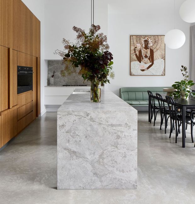 A refined open-plan kitchen and dining space with a large pale natural-stone island, full-height warm timber cabinetry, white cabinetry, and polished concrete flooring. A green upholstered banquette sits beside a dark dining table and chairs, while sculptural pendant lights and artwork add character. The space is bright, contemporary, and softly minimal with natural floral styling.