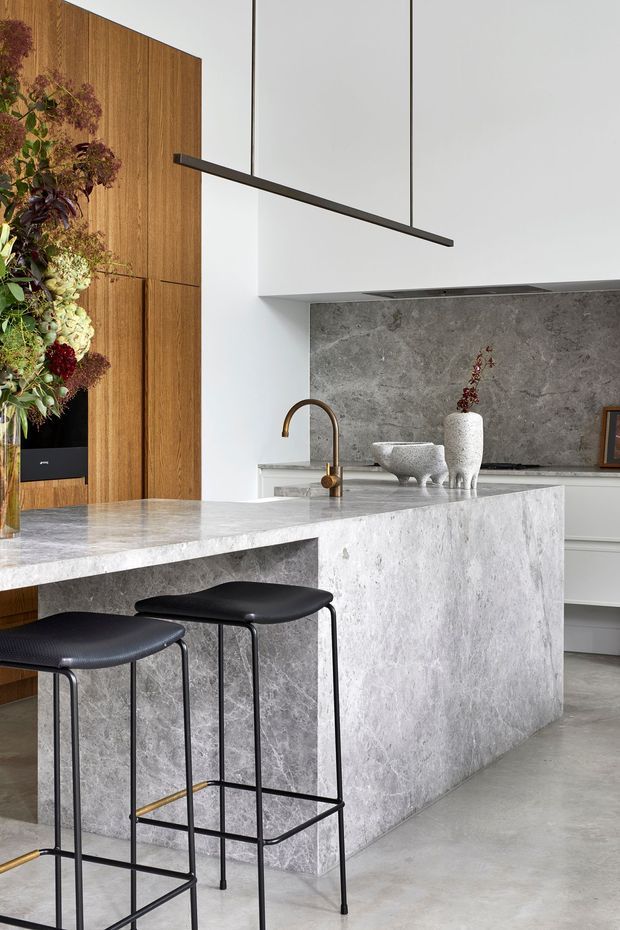 Contemporary kitchen with a large grey stone island and integrated waterfall-style sides, slim black bar stools, a bronze-toned gooseneck tap, white cabinetry, and a matching grey stone splashback. Warm timber wall panelling and concealed appliances contrast with the pale stone and minimalist black linear pendant light.