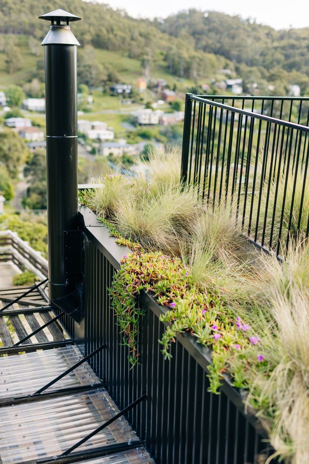 A landscaped elevated outdoor terrace or balcony with black metal safety railings, a translucent metal-grated stairway, and a tall black metal chimney or flue. Built-in planter edges contain ornamental grasses and trailing flowering plants. The terrace overlooks a green, hilly suburban landscape.
