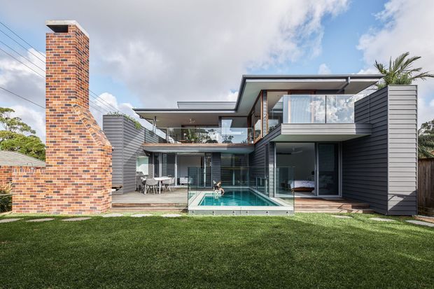 Exterior rear view of a contemporary multi-level residence with dark horizontal cladding, extensive floor-to-ceiling glazing, a projecting upper-level bedroom or living space, and a covered outdoor entertaining area. A narrow swimming pool sits centrally between the house wings, while a prominent exposed red-brick chimney and landscaped lawn occupy the foreground.