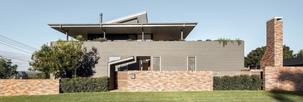 Wide exterior view of a contemporary two-storey house with horizontal grey weatherboard cladding, a broad low-pitched roof with projecting eaves, clerestory-style upper glazing, and a prominent exposed brick chimney. A low brick boundary wall and landscaped hedging frame the front lawn.