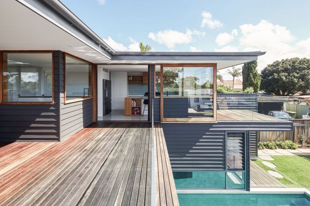 Exterior view of a contemporary, single-storey split-level home with dark horizontal cladding, extensive timber-framed glazing, broad timber decking and a turquoise swimming pool. The flat-roofed building opens onto multiple outdoor decks, with a kitchen visible through the central glazing and landscaped suburban surroundings beyond.