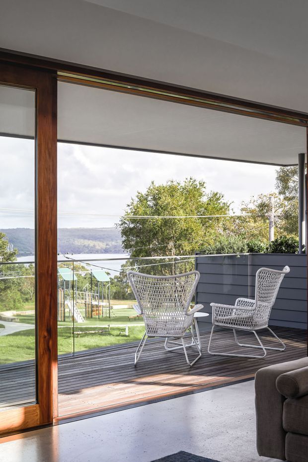 A bright contemporary living room opening onto a raised timber deck through expansive floor-to-ceiling glazing. The deck has glass balustrades, dark horizontal cladding, and two woven outdoor lounge chairs, with a leafy suburban landscape and water view beyond. Inside, a partial upholstered sofa and polished concrete floor are visible; the glazing features warm timber framing and a broad overhead blind or ceiling plane.