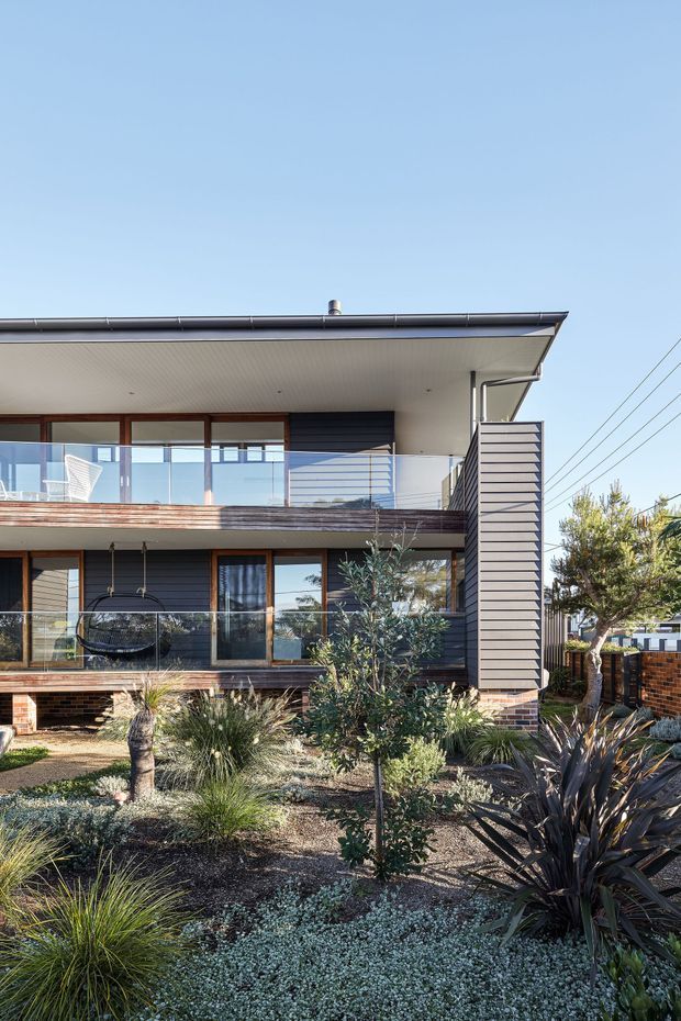 Exterior view of a contemporary two-storey residence with a low-pitched roof, broad upper-level balcony, timber-framed glazing, dark horizontal cladding, brickwork at the lower level, and a landscaped front garden featuring drought-tolerant ornamental planting. The facade has clean horizontal lines and an open outdoor balcony overlooking the garden.