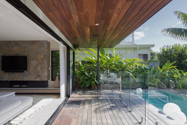 A bright contemporary indoor-outdoor living area opening onto a timber-decked terrace and swimming pool. The covered terrace has a warm timber-lined ceiling, frameless glass balustrade, and timber decking. Inside, a partially visible living room features a large white sofa, stone-clad feature wall, dark built-in media cabinetry, and wide sliding glazing. Tropical planting surrounds the pool.