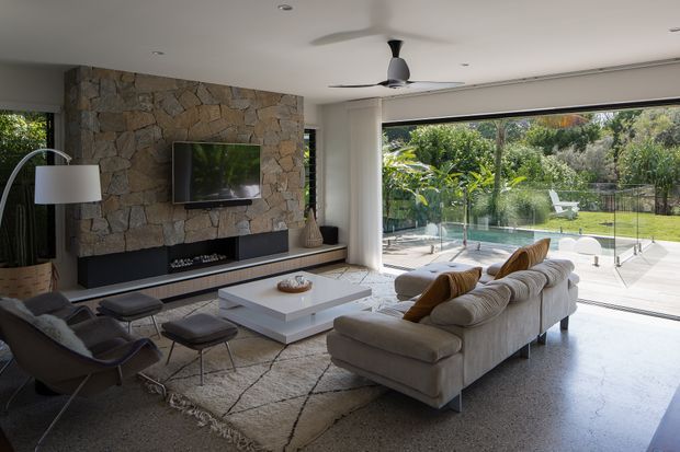 Spacious contemporary living room with a large sectional sofa, lounge chairs, ottomans and a low square coffee table arranged around a wall-mounted television and linear fireplace. A rugged natural-stone feature wall anchors the room, while expansive glazed openings provide views to a landscaped garden and swimming pool. The room has a smooth light floor, recessed ceiling lights and a ceiling fan, with a bright indoor-outdoor connection.
