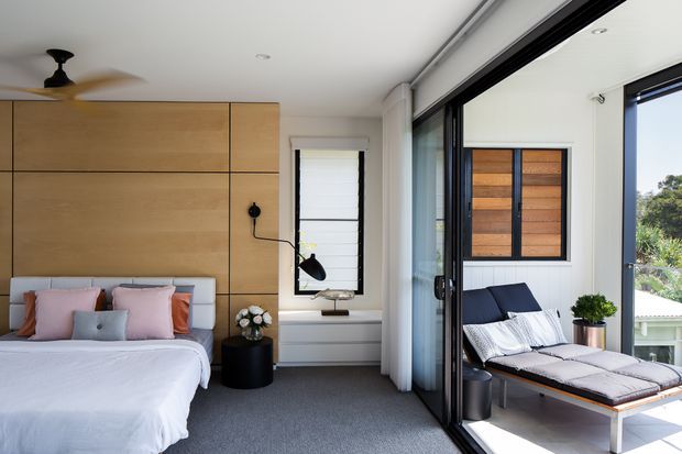 A bright contemporary bedroom with a built-in timber feature wall and integrated storage behind the bed. The room has a grey carpet, white bedding, pastel pink and blue cushions, a black bedside table and lamp, a ceiling fan, and a large black-framed glazed opening leading to an outdoor lounge with a cushioned daybed. A small window with a white roller blind provides additional natural light.