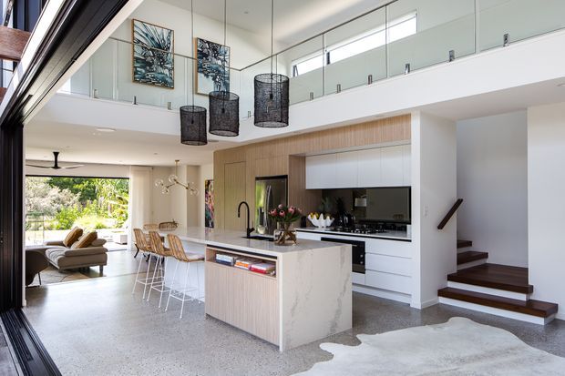 A bright, open-plan contemporary kitchen and living area with a double-height void and mezzanine-level glass balustrade. The kitchen features a large stone island with integrated seating, pale timber cabinetry, white overhead cupboards, and dark appliances. A staircase with dark timber treads leads upward beside the kitchen. The space opens to a leafy outdoor area through large glazed doors, with pendant lighting, polished concrete or terrazzo-like flooring, and neutral furnishings.