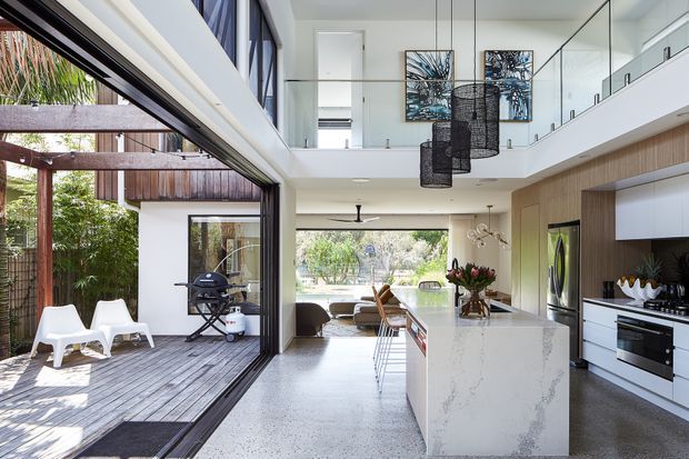 Bright contemporary open-plan kitchen and living space with a double-height volume and mezzanine gallery. A large white stone island with integrated seating anchors the kitchen, which features warm timber cabinetry, white upper cabinets, stainless-steel appliances, and a dark textured floor. Full-height black-framed sliding glass doors open to a timber deck and landscaped outdoor area. Sculptural pendant lights, indoor greenery, and artwork add visual interest.
