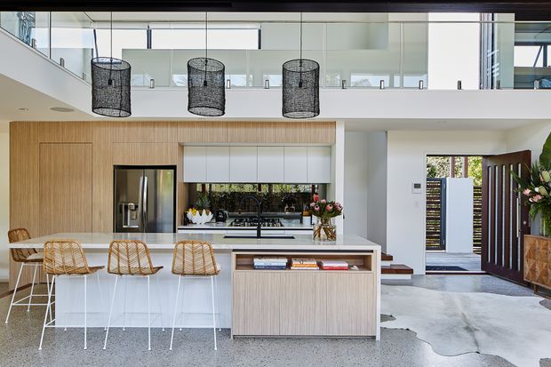 Bright, open-plan contemporary kitchen with a large light-wood island, white stone benchtop, integrated cabinetry, stainless-steel refrigerator, gas cooktop and tiled splashback. Three woven bar stools sit at the island. The space opens toward a timber-framed entry door and includes a double-height area with glass balustrading and three dark woven pendant lights.