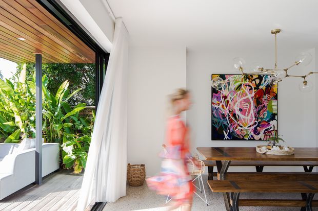 Bright contemporary dining area opening onto a leafy outdoor deck through fully retractable glazed doors. The room has white walls and ceiling, a polished light terrazzo floor, a long solid timber dining table with matching bench seating, a sculptural brass-toned pendant light, and a colourful abstract artwork. A timber-lined outdoor soffit and tropical planting are visible beyond the opening.