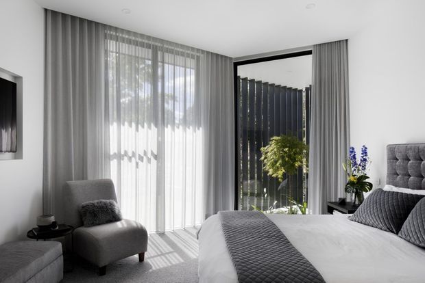 Bright contemporary bedroom with a large upholstered bed, grey quilted bedding, matching grey armchair, floor-to-ceiling sheer curtains, and full-height glazing opening toward a planted outdoor area. The palette is predominantly soft white and grey with dark window framing and subtle greenery accents.