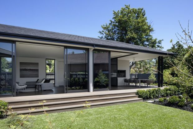 A contemporary single-storey pavilion-style home with a low-pitched dark roof, extensive sliding glass walls, and a broad covered outdoor entertaining area. The open interior includes a living space and outdoor dining area, connected by a raised timber deck and steps to a landscaped lawn and garden.