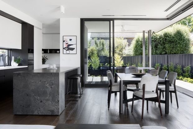 Bright contemporary open-plan kitchen and dining area with a large dark stone island, white cabinetry, dark timber flooring, and a formal dining table beside expansive black-framed sliding glass doors opening to a landscaped outdoor area.