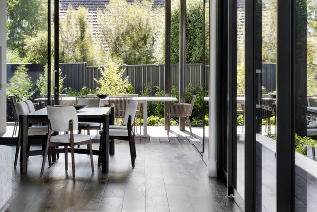A bright contemporary dining area opening onto a landscaped outdoor terrace through extensive black-framed glazing. The interior features a dark timber dining table with upholstered chairs, pale timber flooring, and a slim outdoor dining setting beyond. Reflections in the glazing extend the view of the furnished space and garden.